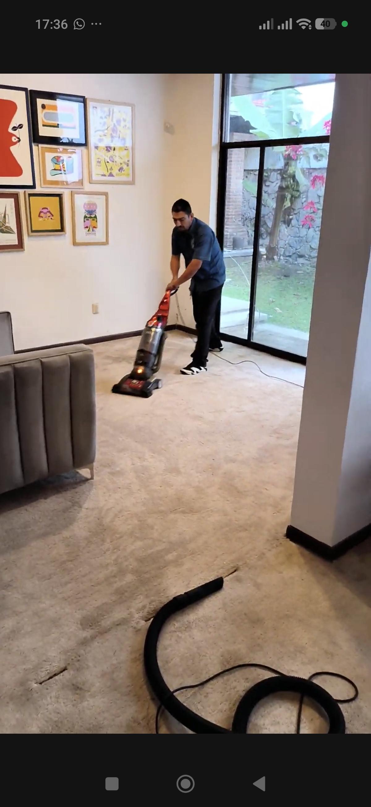 Persona aspirando alfombra en sala con ventanales y sillón gris.