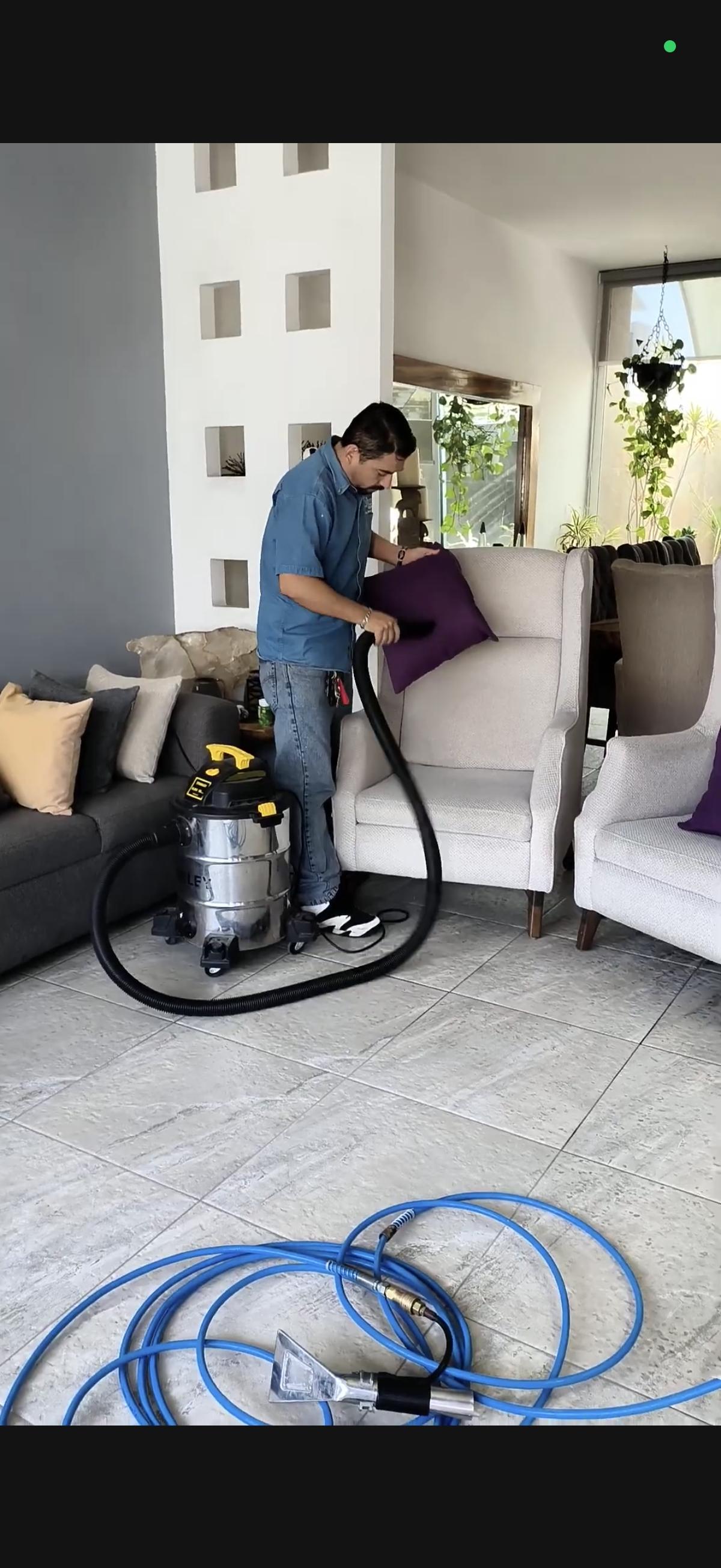 Persona usando aspiradora en sillón blanco junto a sofá gris en una sala iluminada.
